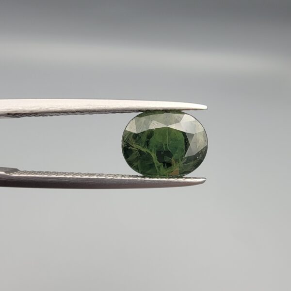 a premium, beautifully faceted 3.12-carat natural Green Sapphire gemstone is held securely in metal tweezers against a clean, neutral grey background. The loose corundum gem showcases an expert oval cut with a well-proportioned table, displaying a rich, uniform olive-green color with fine, genuine internal crystal characteristics that verify its earth-mined origin.