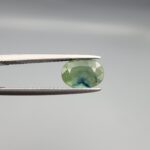 a premium, beautifully faceted 2.12-carat natural Green Sapphire gemstone is held securely in metal tweezers against a clean, neutral grey background. The loose corundum gem showcases an expert oval cut with a well-proportioned table, displaying a rich, uniform olive-green color with a unique blue color-zone patch at its center and fine, genuine internal crystal characteristics that verify its earth-mined origin.