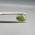 ​A premium, beautifully faceted 2.23-carat natural Green Sapphire gemstone is held securely in metal tweezers against a clean, neutral grey background. The loose corundum gem showcases an expert oval cut with a well-proportioned table, displaying a rich, uniform olive-green color with fine, genuine internal crystal characteristics that verify its earth-mined origin.