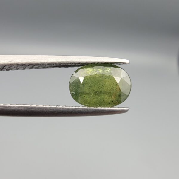 a premium, beautifully faceted 1.58-carat natural Green Sapphire gemstone is held securely in metal tweezers against a clean, neutral grey background. The loose corundum gem showcases an expert oval cut with a well-proportioned table, displaying a rich, uniform olive-green color with fine, genuine internal crystal characteristics that verify its earth-mined origin.