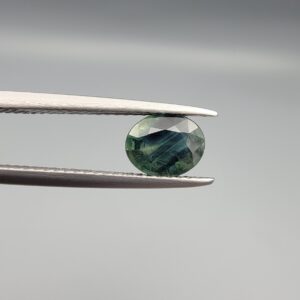a premium, beautifully faceted 1.45-carat natural Green Sapphire gemstone is held securely in metal tweezers against a clean, neutral grey background. The loose corundum gem showcases an expert oval cut with a well-proportioned table, displaying a rich, deep olive-green color with fascinating natural internal silk patterns and genuine crystal characteristics that verify its earth-mined origin.