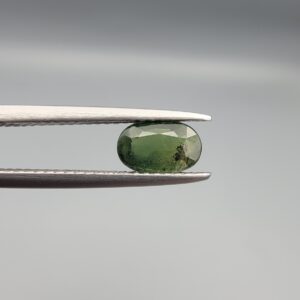 a premium, beautifully faceted 1.06-carat natural Green Sapphire gemstone is held securely in metal tweezers against a clean, neutral grey background. The loose corundum gem showcases an expert oval cut with a well-proportioned table, displaying a rich, uniform olive-green color with fine, genuine internal crystal characteristics that verify its earth-mined origin.