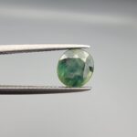 a premium, beautifully faceted 2.27-carat natural Green Sapphire gemstone is held securely in metal tweezers against a clean, neutral grey background. The loose corundum gem showcases an expert oval cut with a well-proportioned table, displaying a rich olive-green body color with distinct, genuine internal blue-green color bands and natural crystal characteristics that verify its earth-mined origin.