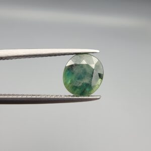 a premium, beautifully faceted 2.27-carat natural Green Sapphire gemstone is held securely in metal tweezers against a clean, neutral grey background. The loose corundum gem showcases an expert oval cut with a well-proportioned table, displaying a rich olive-green body color with distinct, genuine internal blue-green color bands and natural crystal characteristics that verify its earth-mined origin.