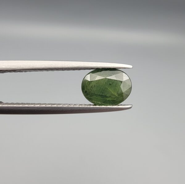 a premium, beautifully faceted 1.42-carat natural Green Sapphire gemstone is held securely in metal tweezers against a clean, neutral grey background. The loose corundum gem showcases an expert oval cut with a well-proportioned table, displaying a rich, uniform olive-green color with fine, genuine internal crystal characteristics that verify its earth-mined origin.