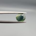 ​A premium, beautifully faceted 1.20-carat natural Green Sapphire gemstone is held securely in metal tweezers against a clean, neutral grey background. The loose corundum gem showcases an expert oval cut with a well-proportioned table, displaying a rich olive-green body color with a distinct, genuine internal deep blue color-zone patch and natural crystal characteristics that verify its earth-mined origin.