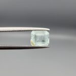 A 1.20-carat natural Pakistani Aquamarine gemstone held horizontally in professional metal gemstone tweezers against a smooth gray studio background. The loose beryl gem is fashioned into an octagon step cut, showcasing a pale, translucent ice-blue to pastel-aqua body color filled with fine internal growth silk and unique organic mountain inclusions near its bottom corner.