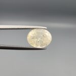 A premium, high-clarity 2.67-carat natural Ceylon Yellow Sapphire gemstone held horizontally in professional steel gemstone tweezers against a smooth gray studio background. The loose Sri Lankan Pukhraj features an elegant faceted oval cut, displaying a vibrant, evenly distributed lemon-yellow color profile with immaculate crown facet symmetry and intense internal light refraction.