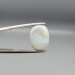 ​A prominent 5.19-carat natural Australian Opal gemstone held firmly in professional metal gemstone tweezers against a smooth grey studio background. The loose oval cabochon exhibits a smooth, polished dome surface reflecting vibrant neon flashes of color, while its reverse side features completely organic, naturally formed mountain fissures, minor cracks, and host rock holes that verify its raw earth-mined authenticity.