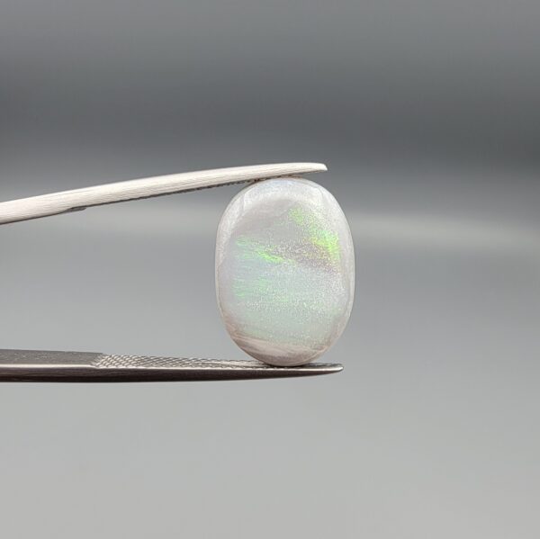 ​A prominent 5.19-carat natural Australian Opal gemstone held firmly in professional metal gemstone tweezers against a smooth grey studio background. The loose oval cabochon exhibits a smooth, polished dome surface reflecting vibrant neon flashes of color, while its reverse side features completely organic, naturally formed mountain fissures, minor cracks, and host rock holes that verify its raw earth-mined authenticity.