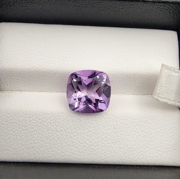 ​A high-clarity 4.21-carat natural Brazilian Amethyst gemstone beautifully presented in a luxury white-cushioned black leatherette display box. The loose gem showcases a brilliant cushion cut with softly rounded rectangular edges, displaying a uniform, bright violet-purple color profile with magnificent internal light performance and sharp facet symmetry.