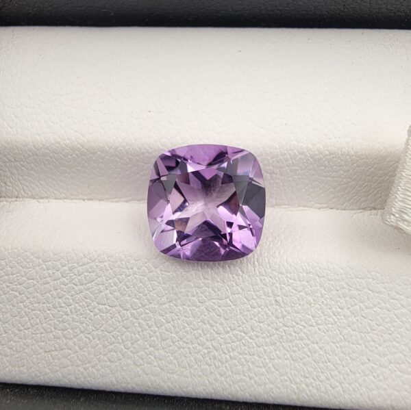 ​A high-clarity 4.21-carat natural Brazilian Amethyst gemstone beautifully presented in a luxury white-cushioned black leatherette display box. The loose gem showcases a brilliant cushion cut with softly rounded rectangular edges, displaying a uniform, bright violet-purple color profile with magnificent internal light performance and sharp facet symmetry.