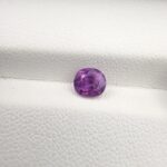 A premium, high-clarity 1.04-carat natural Ceylon Purple Sapphire gemstone placed on a white-textured leatherette background. The loose gem showcases an elegant faceted cushion cut, displaying an intensely saturated, deep royal purple and vibrant violet body color with rich internal facet complexity and sharp light refraction.