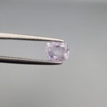 A premium, high-clarity 1.08-carat natural Ceylon Light Purple Sapphire gemstone held securely in metal tweezers against a clean grey background. The loose gem showcases an elegant faceted cushion cut with beautifully balanced crown facets, displaying a delicate, brightly translucent light violet-purple body color with excellent internal light reflection and pristine facet symmetry.