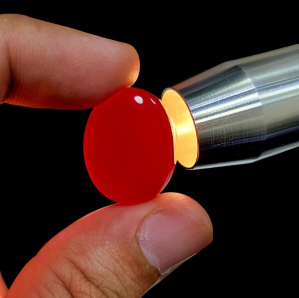​A perfectly polished 41.56-carat natural oval Yemeni Carnelian Agate cabochon held securely between fingers against a sleek grey background, showing off a glossy liver-brown (Kaleji) surface color. A secondary view showcases the gemstone nestled elegantly inside a premium black and white cushioned display box. A third close-up view demonstrates the stone being backlit directly by a professional gem torch, causing the entire oval cabochon to radiate a vibrant, translucent fiery red (Laal) from within its flawless internal structure.