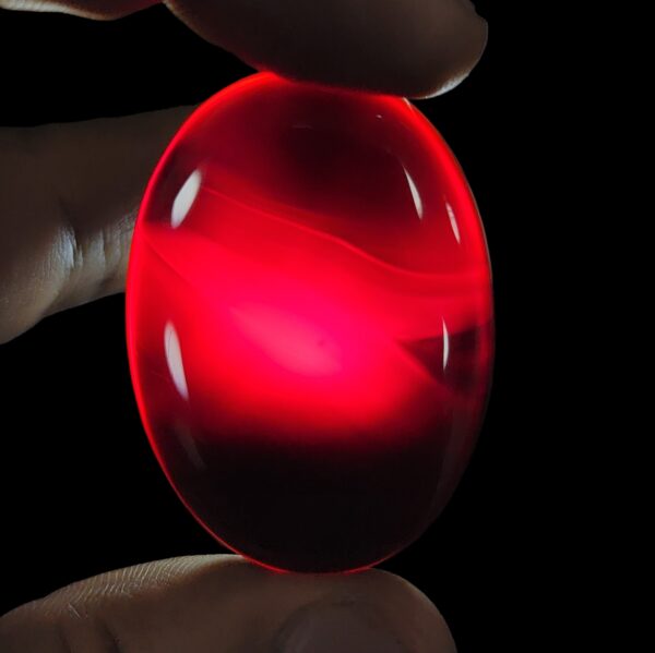 A giant, masterfully polished 85.42-carat natural oval Yemeni Carnelian Agate cabochon is held between fingers against a grey background, displaying a flawless mirror-like gloss and a rich, deep liver-brown (Kaleji) color. In a secondary view under dark conditions, the exact same gemstone is illuminated by a torch light from behind, revealing a brilliant, glowing translucent red (Laal) interior adorned with delicate, fine natural agate structural banding.