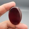 A giant, masterfully polished 85.42-carat natural oval Yemeni Carnelian Agate cabochon is held between fingers against a grey background, displaying a flawless mirror-like gloss and a rich, deep liver-brown (Kaleji) color. In a secondary view under dark conditions, the exact same gemstone is illuminated by a torch light from behind, revealing a brilliant, glowing translucent red (Laal) interior adorned with delicate, fine natural agate structural banding.