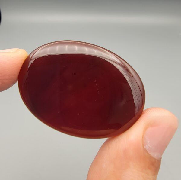 A giant, masterfully polished 85.42-carat natural oval Yemeni Carnelian Agate cabochon is held between fingers against a grey background, displaying a flawless mirror-like gloss and a rich, deep liver-brown (Kaleji) color. In a secondary view under dark conditions, the exact same gemstone is illuminated by a torch light from behind, revealing a brilliant, glowing translucent red (Laal) interior adorned with delicate, fine natural agate structural banding.