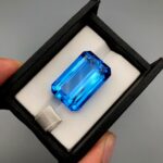 ​A massive, high-clarity 46.42-carat natural Swiss Blue Topaz gemstone presented inside a professional black leatherette display case with a white cushioned interior. The loose, earth-mined gem is cut into a flawless rectangular emerald-style shape, displaying an intense, vivid electric-blue body color with perfect parallel step facets and immaculate crown reflections.