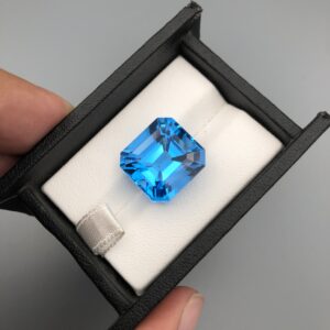 ​A premium, high-clarity 23.04-carat natural Swiss Blue Topaz gemstone centered inside a professional black leatherette display box with a white cushioned base. The loose, earth-mined gem is meticulously fashioned into a square Asscher cut, exhibiting a brilliant, electric-blue body color with perfect concentric step facets and intense internal light refraction.