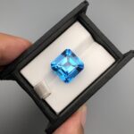 ​A premium, highly translucent 20.20-carat natural Swiss Blue Topaz gemstone displayed face-up inside a professional black leatherette display box lined with white textured fabric. The loose gem features a crisp, square Asscher cut with cropped corners, showcasing an even, vibrant electric-blue hue with sharp step facets and dazzling internal reflections.