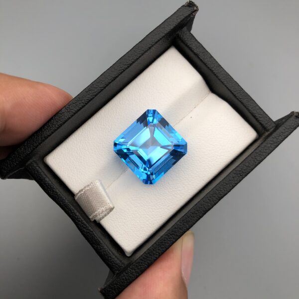 ​A premium, highly translucent 20.20-carat natural Swiss Blue Topaz gemstone displayed face-up inside a professional black leatherette display box lined with white textured fabric. The loose gem features a crisp, square Asscher cut with cropped corners, showcasing an even, vibrant electric-blue hue with sharp step facets and dazzling internal reflections.