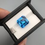 A premium, exceptionally clear 18.31-carat natural Swiss Blue Topaz gemstone presented face-up inside a professional black leatherette display box with a white textured fabric interior. The loose gem features a precision-cut square Asscher shape with elegant cropped corners, showcasing an even, vibrant electric-blue color with sharp concentric step facets.