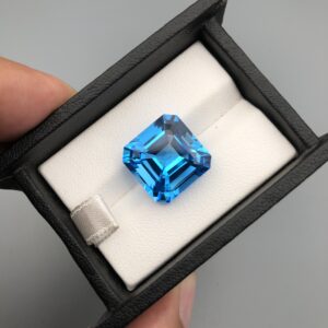 A premium, exceptionally clear 18.31-carat natural Swiss Blue Topaz gemstone presented face-up inside a professional black leatherette display box with a white textured fabric interior. The loose gem features a precision-cut square Asscher shape with elegant cropped corners, showcasing an even, vibrant electric-blue color with sharp concentric step facets.