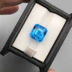 A premium, exceptionally clear 16.39-carat natural Swiss Blue Topaz gemstone presented face-up inside a professional black leatherette display box with a white textured fabric interior. The loose gem features a precision square Asscher shape with elegant cropped corners, showcasing an even, vibrant electric-blue color with sharp concentric step facets and beautiful internal light performance.
