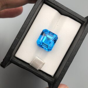 A premium, exceptionally clear 16.39-carat natural Swiss Blue Topaz gemstone presented face-up inside a professional black leatherette display box with a white textured fabric interior. The loose gem features a precision square Asscher shape with elegant cropped corners, showcasing an even, vibrant electric-blue color with sharp concentric step facets and beautiful internal light performance.
