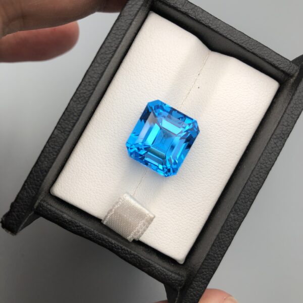 A premium, exceptionally clear 16.39-carat natural Swiss Blue Topaz gemstone presented face-up inside a professional black leatherette display box with a white textured fabric interior. The loose gem features a precision square Asscher shape with elegant cropped corners, showcasing an even, vibrant electric-blue color with sharp concentric step facets and beautiful internal light performance.
