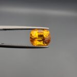 a premium, beautifully faceted 4.45-carat natural Vivid Orangish Yellow Sapphire gemstone is held securely in metal tweezers against a clean, neutral grey background. The loose cushion-cut corundum gem displays magnificent clarity and a highly saturated, rich golden-orange color with intense internal fire and brilliant light reflection.
