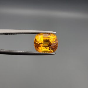 a premium, beautifully faceted 4.45-carat natural Vivid Orangish Yellow Sapphire gemstone is held securely in metal tweezers against a clean, neutral grey background. The loose cushion-cut corundum gem displays magnificent clarity and a highly saturated, rich golden-orange color with intense internal fire and brilliant light reflection.
