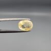 a premium, beautifully faceted 3.22-carat natural Yellow Sapphire gemstone is held securely in metal tweezers against a clean, neutral grey background. The loose corundum gem showcases an expert oval cut with a well-proportioned table, displaying a bright, light-yellow color with fine, genuine internal crystal characteristics and natural feather inclusions that verify its rare, unheated earth-mined origin.