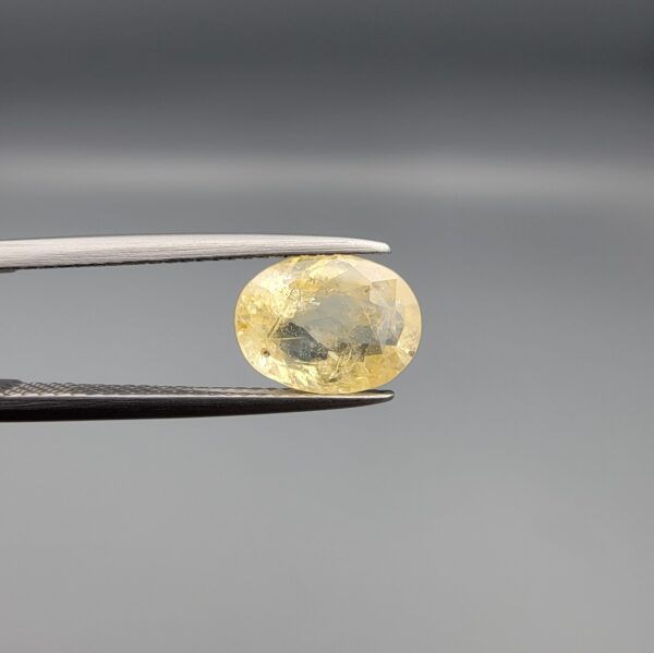 a premium, beautifully faceted 3.22-carat natural Yellow Sapphire gemstone is held securely in metal tweezers against a clean, neutral grey background. The loose corundum gem showcases an expert oval cut with a well-proportioned table, displaying a bright, light-yellow color with fine, genuine internal crystal characteristics and natural feather inclusions that verify its rare, unheated earth-mined origin.