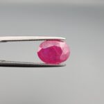 ​A 3.13-carat oval faceted natural Mozambique ruby held in metallic tweezers against a neutral gray background, displaying a bright purplish-red hue and visible internal silk inclusions.