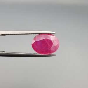 ​A 3.13-carat oval faceted natural Mozambique ruby held in metallic tweezers against a neutral gray background, displaying a bright purplish-red hue and visible internal silk inclusions.