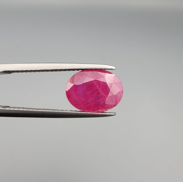 ​A 3.13-carat oval faceted natural Mozambique ruby held in metallic tweezers against a neutral gray background, displaying a bright purplish-red hue and visible internal silk inclusions.