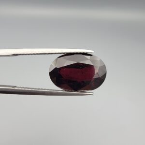 A premium, beautifully faceted 8.31-carat natural Red Garnet gemstone is held securely in metal tweezers against a clean, neutral grey background. The loose gemstone showcases an expert oval cut with a well-proportioned table, displaying a deep, rich crimson-red color with fine, genuine internal characteristics that verify its authentic earth-mined origin.