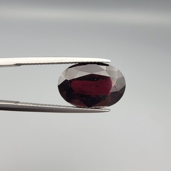 ​A premium, beautifully faceted 8.31-carat natural Red Garnet gemstone is held securely in metal tweezers against a clean, neutral grey background. The loose gemstone showcases an expert oval cut with a well-proportioned table, displaying a deep, rich crimson-red color with fine, genuine internal characteristics that verify its authentic earth-mined origin.