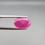 ​A premium, beautifully faceted 3.50-carat natural Ruby gemstone is held securely in metal tweezers against a clean, neutral grey background. The loose corundum gem showcases an expert oval cut with a well-proportioned table, displaying a vibrant pinkish-red color with fine, genuine internal crystal characteristics and natural silk inclusions that verify its rare, unheated earth-mined origin.