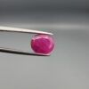 ​A premium, beautifully faceted 3.35-carat natural Ruby gemstone is held securely in metal tweezers against a clean, neutral grey background. The loose corundum gem showcases an expert oval cut with a well-proportioned table, displaying a rich pinkish-red color with fine, genuine internal crystal characteristics and natural silk inclusions that verify its rare, unheated earth-mined origin.