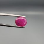 ​A premium, beautifully faceted 3.35-carat natural Ruby gemstone is held securely in metal tweezers against a clean, neutral grey background. The loose corundum gem showcases an expert oval cut with a well-proportioned table, displaying a rich pinkish-red color with fine, genuine internal crystal characteristics and natural silk inclusions that verify its rare, unheated earth-mined origin.