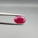 A premium, beautifully faceted 2.17-carat natural Ruby gemstone is held securely in metal tweezers against a clean, neutral grey background. The loose corundum gem showcases an expert oval cut with a well-proportioned table, displaying a rich, vibrant red color with fine, genuine internal crystal characteristics and natural silk inclusions that verify its rare, unheated earth-mined origin.