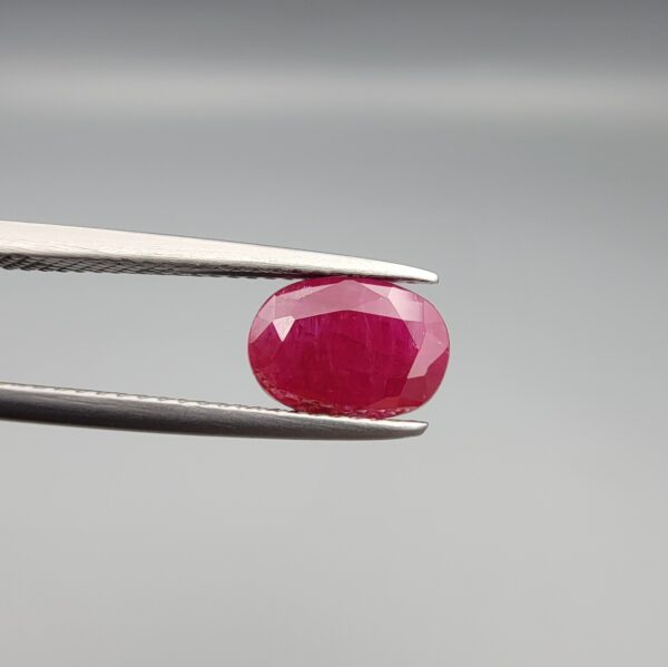 A premium, beautifully faceted 2.17-carat natural Ruby gemstone is held securely in metal tweezers against a clean, neutral grey background. The loose corundum gem showcases an expert oval cut with a well-proportioned table, displaying a rich, vibrant red color with fine, genuine internal crystal characteristics and natural silk inclusions that verify its rare, unheated earth-mined origin.