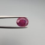 ​A premium, beautifully faceted 2.27-carat natural Ruby gemstone is held securely in metal tweezers against a clean, neutral grey background. The loose corundum gem showcases an expert oval cut with a well-proportioned table, displaying a rich, vibrant purplish-red color with fine, genuine internal crystal characteristics and natural silk inclusions that verify its rare, unheated earth-mined origin.