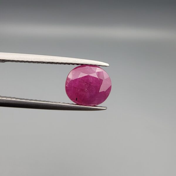 ​A premium, beautifully faceted 2.68-carat natural Ruby gemstone is held securely in metal tweezers against a clean, neutral grey background. The loose corundum gem showcases an expert oval cut with a well-proportioned table, displaying a rich red color with fine, genuine internal crystal characteristics and natural silk inclusions that verify its rare, unheated earth-mined origin.