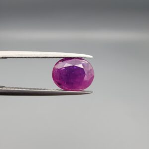 A premium, beautifully faceted 2.68-carat natural Ruby gemstone is held securely in metal tweezers against a clean, neutral grey background. The loose corundum gem showcases an expert oval cut with a well-proportioned table, displaying a rich, vibrant purplish-red color with fine, genuine internal crystal characteristics and natural silk inclusions that verify its rare, unheated earth-mined origin.