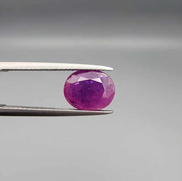 A premium, beautifully faceted 2.68-carat natural Ruby gemstone is held securely in metal tweezers against a clean, neutral grey background. The loose corundum gem showcases an expert oval cut with a well-proportioned table, displaying a rich, vibrant purplish-red color with fine, genuine internal crystal characteristics and natural silk inclusions that verify its rare, unheated earth-mined origin.
