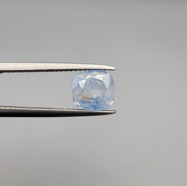 a premium, beautifully faceted 2.95-carat natural Blue Sapphire gemstone is held securely in metal tweezers against a clean, neutral grey background. The loose corundum gem showcases an expert cushion cut with a well-proportioned table, displaying an icy light-blue color with fine, genuine internal crystal characteristics and natural feather inclusions that verify its rare, unheated earth-mined origin.